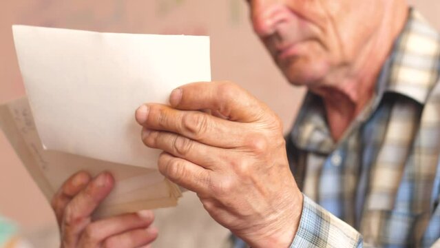Old archival photos are in the wrinkled hands of an elderly man. A 70-79-year-old Caucasian pensioner examines family photos. Selective focus.Slow motion