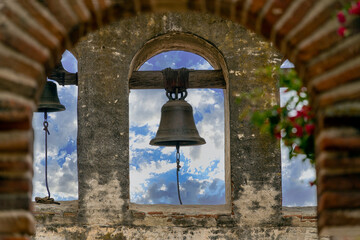 Historic church bells hanging in stone archway against a blue cloudy sky outside. 