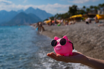 A woman holds a piggy bank in sunglasses on a pebble beach near the sea. Budget vacation.