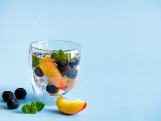 Blueberry and peach infused water, cocktail, lemonade or tea in double glass. Summer iced cold drink with blueberry, ice, peach and mint on blue paper background, copy space, side view