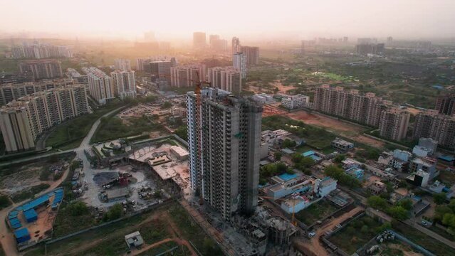aerial orbiting shot of under construction skyscraper with crane and workers with parallax effect with the smaller houses and buildings in the background shot at dusk in the city of gurgaon delhi