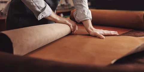 Deurstickers Cobbler working with skin textile in workshop. Tailor hold different rolls natural brown leather © Parilov
