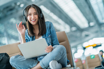 attractive asian female traveller wear casula cloth headphone using laptop for wating flight departure at airport terminal,asian female smile hand waving greeting looking at camera travel idea concept