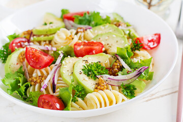 Fusilli pasta salad with avocado, tomatoes, fresh green lettuce, red onion and mustard dressing on white background. Vegetarian healthy lunch.