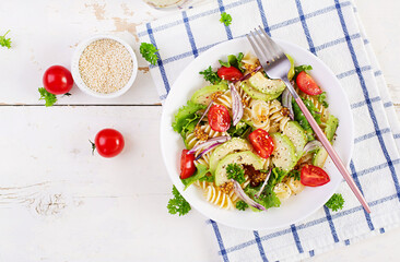 Fusilli pasta salad with avocado, tomatoes, fresh green lettuce, red onion and mustard dressing on white background. Vegetarian healthy lunch. Top view, flat lay