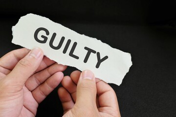 Guilty verdict concept. Hand holding a piece of paper with written word guilty in black background.