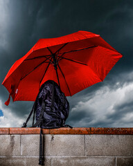 A black backpack protected by a red umbrella against a cloudy sky that threatens rain
