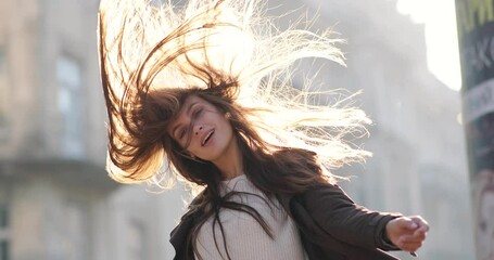 Attractive brunette woman has fun. She is staying in the street and shaking her head, playing with hair and looking at the camera. The wind is blowing and her hair fluttering. The rays of sun shines