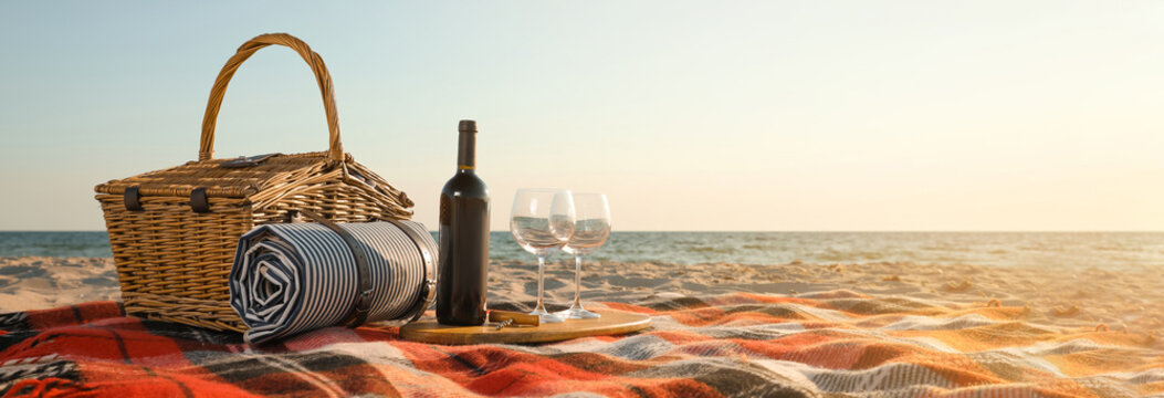 Blanket with picnic basket, bottle of wine and glasses on sandy beach near sea, space for text. Banner design