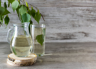 Birch sap in a transparent jug on a wooden stand. A tall glass of sap and a sprig of birch in the background. Light wooden background, space for text