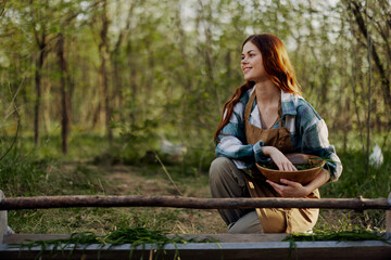 Naklejka premium A girl bird farm worker smiles and is happy pouring food into the chicken feeder in the fresh air sitting on the green grass
