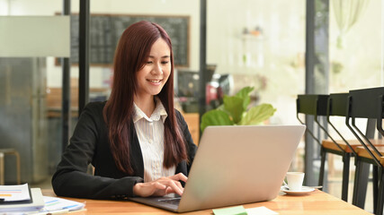 Fototapeta premium Smiling asian woman in business suit using laptop computer at coffee shop