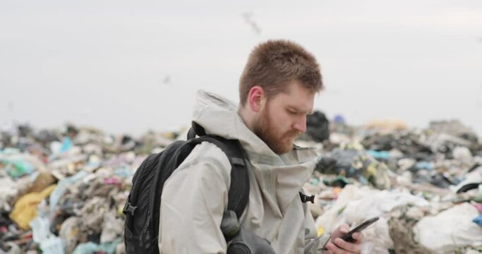 A working man against the backdrop of burning garbage. A lot of plastic bags thrown to the dump. From the plastic debris goes smoke. On the man, the khaki color encephalic suit