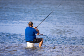 An unidentifiable middle aged man with a fishing rod sits on a bucket in a shallow lake, surrounded by calm blue water, on a sunny afternoon. Copy space. - Powered by Adobe