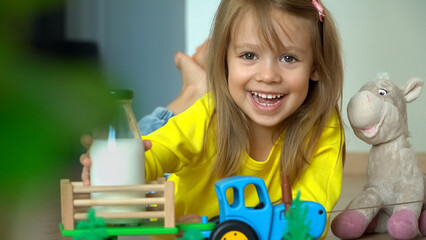 Portrait of little girl with milk bottle looks at camera and laughs. Toy tractor delivers milk in cart to happy child. Drink milk