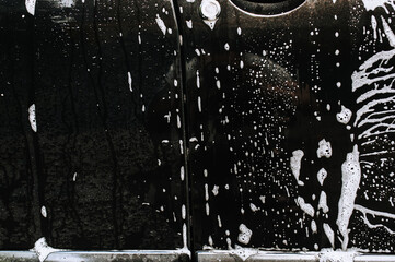 Background, texture of the black surface of a metal car door in white soapy foam after washing. Photography, concept of shampoo streaks.