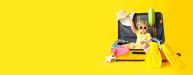 Adorable baby girl sitting in big suitcase with belongings on yellow background with space for text