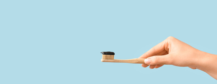 Female hand holding bamboo brush with activated charcoal toothpaste on light blue background with space for text