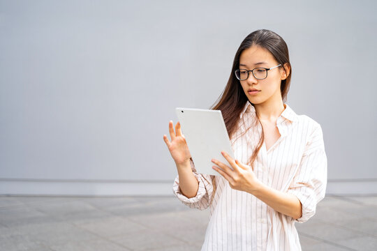 Young Asian Thai, Vietnamese or Chinese woman in shirt and glasses standing outside near office using modern tablet with webcam reading working, learning or chatting. Education, wireless technilogy