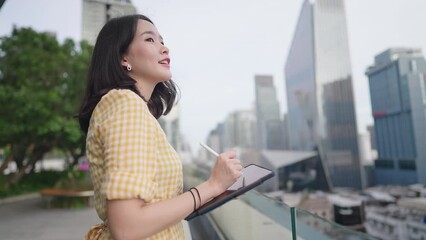 Smart Asian girl using tablet and smartpen thinking of new creative ideas while finding a new work inspiration, woman standing in the middle of city business area, thoughtful hard working woman
