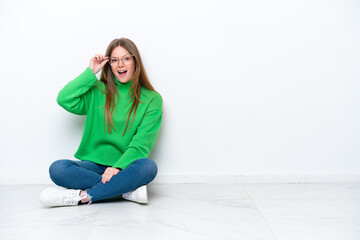 Young caucasian woman sitting on the floor isolated on white background with glasses and surprised