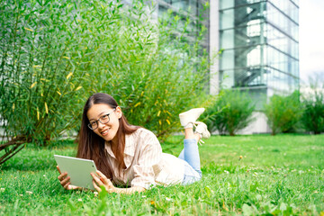 Portrait of young smiling Asian Thai, Vietnamese or Chinese woman in casual clothes and glasses lying on green grass loan outside near univercity or school working reading or learning using tablet