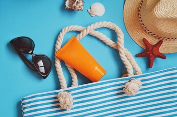 Flat lay summer travel lifestyle photo. Stylish sunglasses, straw hat, sunscreen cream, striped bag and seashells on a blue background. Beach essentials, travelling pack top view