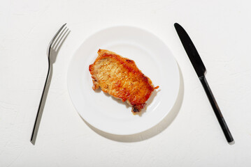 A piece of fried meat on a white plate. Black knife and fork. Bad fatty food. White background. Diet keto concept.