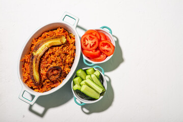 Jollof rice with fried banana. Fresh vegetables - tomato and cucumber. White background. Traditional Nigerian rice food with tomatoes, onions and spices.