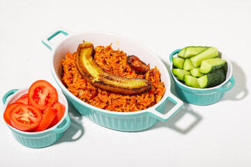 Jollof rice with fried banana. Fresh vegetables - tomato and cucumber. White background. Traditional Nigerian rice food with tomatoes, onions and spices.