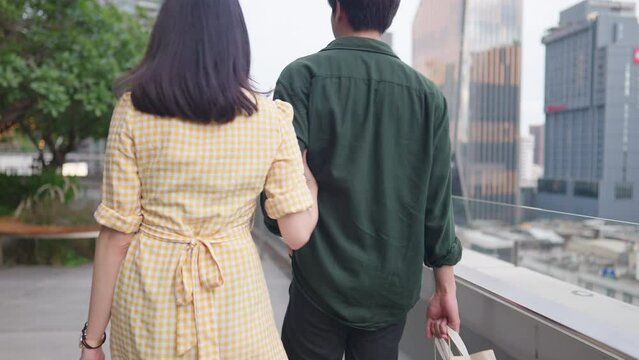 Young adult Asian couple walking along city pedestrian outdoor garden, woman hold on to boyfriend arms, trust reliable love romantic relationship, credit card member point redemption privileges