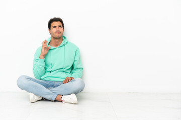 Caucasian handsome man sitting on the floor with fingers crossing and wishing the best