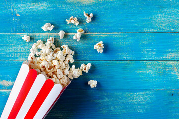Fresh popcorn spilled from a cardboard striped cup on a blue wooden background with copy space. Top...