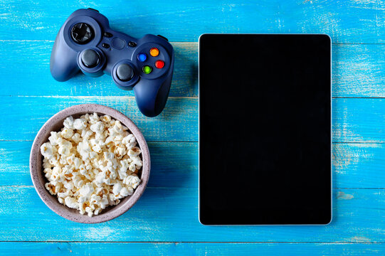 Bowl of popcorn, game console and tablet on blue wooden background with copy space. Top view. The concept of leisure, games, pastime.