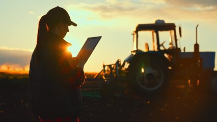 Farmer woman with tablet working in field. Concept of technology in agriculture. Harvest season in an organic farm on a bright sunny day. Tractor at work. - Powered by Adobe