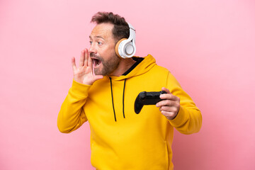 Middle age man playing with a video game controller isolated on pink background shouting with mouth wide open to the side