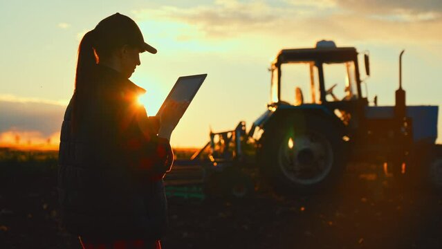 Farmer woman with tablet working in field. Concept of technology in agriculture. Harvest season in an organic farm on a bright sunny day. Tractor at work.
