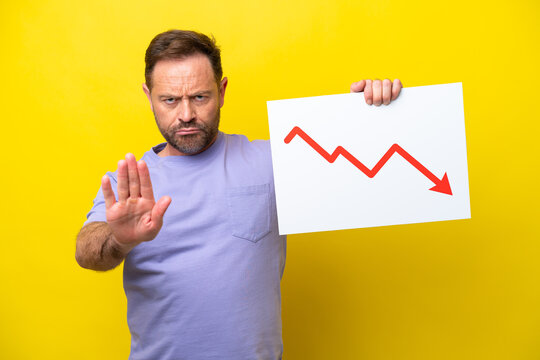 Middle age caucasian man isolated on yellow background holding a sign with a decreasing statistics arrow symbol and doing stop sign
