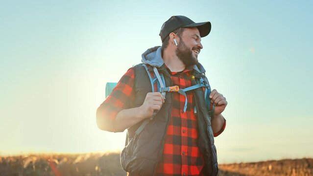 Man traveler walks through field with backpack on his shoulders. Tourist route for spending time in beautiful nature. Outdoor landscape with plants on ground. Active lifestyle on move. Sun rays.