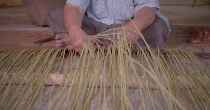 Cut in shot of an Asian female farmer who is an elderly native basketry artisan weave mat from bamboo that are cut into strips for weaving folk crafts in rural areas of Thailand, southeast Asia.
