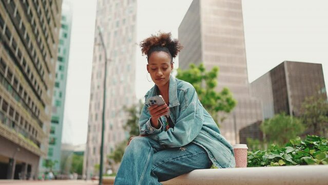 Cute African girl with ponytail, wearing denim jacket with disposable coffee cup sits and looks through photos, videos on mobile phone on modern buildings background. Slow motion