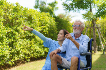 Asian caregiver nurse with senior man sitting on wheelchair in garden and looking nature around home in morning.