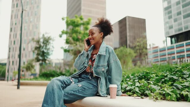 Beautiful African girl with ponytail, wearing denim jacket, in crop top with national pattern talking on mobile phone on modern city background. Slow motion