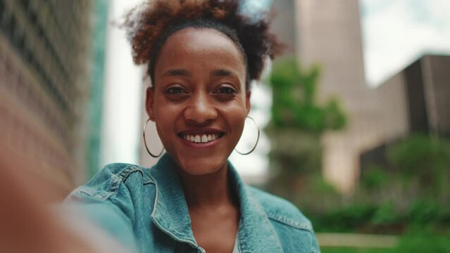 Closeup portret cute African girl with ponytail, wearing denim jacket, sits with morning coffee and makes stream, video call on modern city background. Slow motion