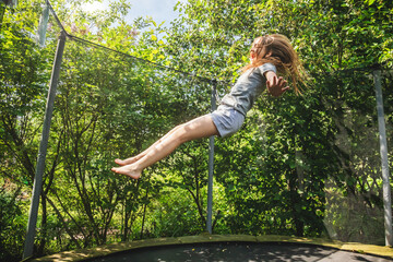 Preteen girl having fun bouncing on a trampoline in a backyard on a summer day - obrazy, fototapety, plakaty