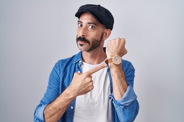 Hispanic man with beard standing over isolated background in hurry pointing to watch time,...