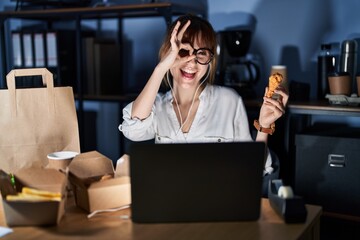 Obraz premium Young beautiful woman working using computer laptop and eating delivery food doing ok gesture with hand smiling, eye looking through fingers with happy face.