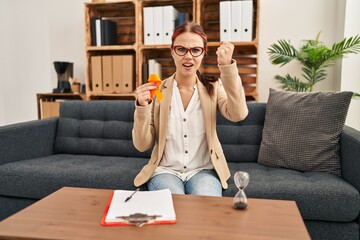 Young caucasian woman holding awareness orange ribbon at the office annoyed and frustrated shouting...