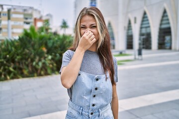Young caucasian woman outdoors smelling something stinky and disgusting, intolerable smell, holding breath with fingers on nose. bad smell - obrazy, fototapety, plakaty