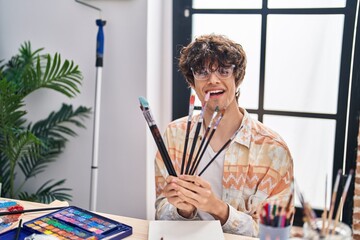Young hispanic man holding paintbrushes sitting on desk at art studio